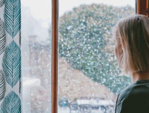 blonde woman looking out the window of her home with snow in the background